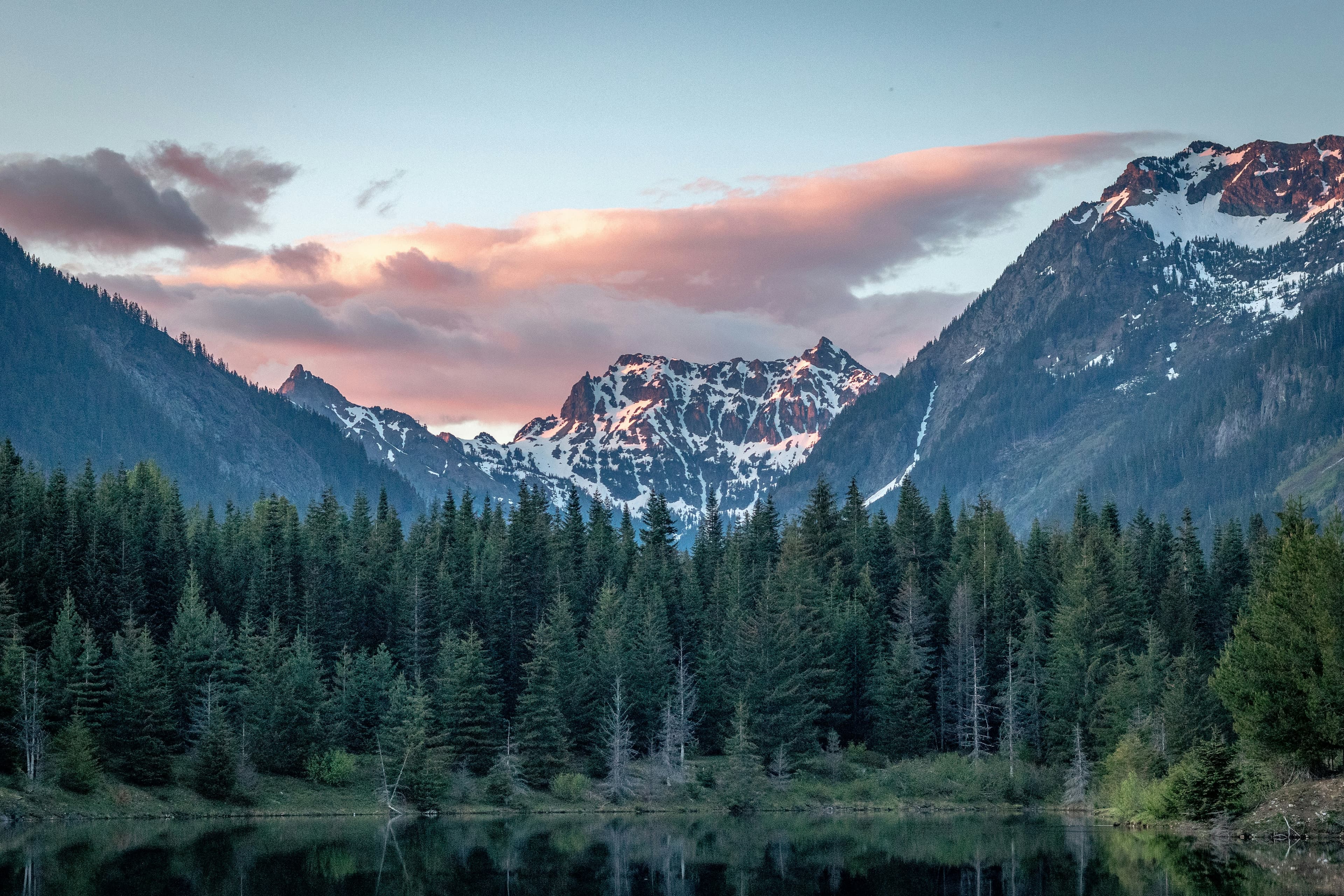 A mountain lake with pine trees and snow-covered peaks at sunset.